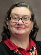 A woman with glasses and shoulder-length hair smiles. She wears a red and black outfit with a patterned scarf and pearl earrings against a plain background.