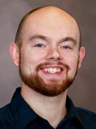 A man with a bald head, neatly trimmed beard, and mustache is smiling. He is wearing a dark shirt and looking directly at the camera against a neutral background.